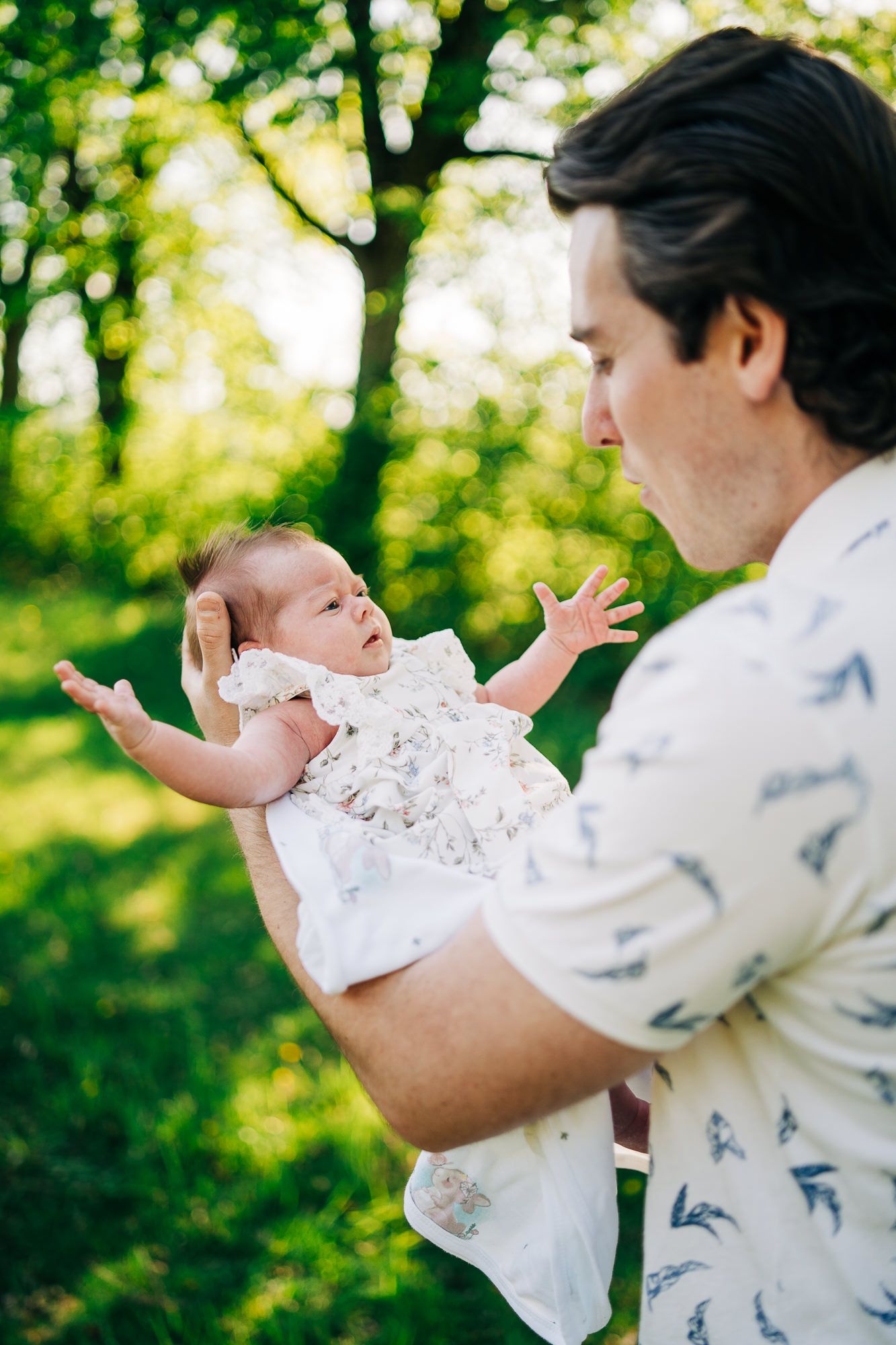 A father in a printed white shirt holds a small baby in a white dress outdoors with green trees in the background.