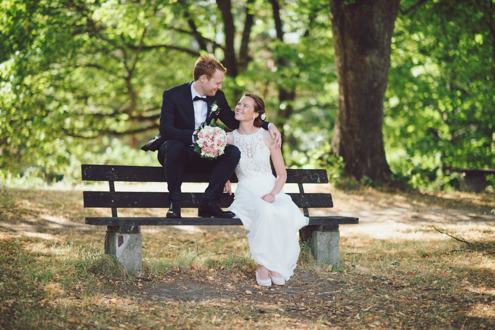 Wedding couple shares an intimate laugh while sitting on a wooden bench in a sun-dappled forest.