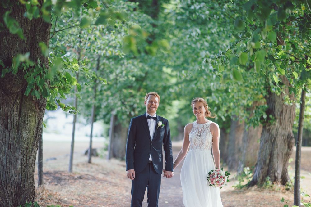 Newlyweds hold hands while walking through tree-lined path in elegant wedding attire.