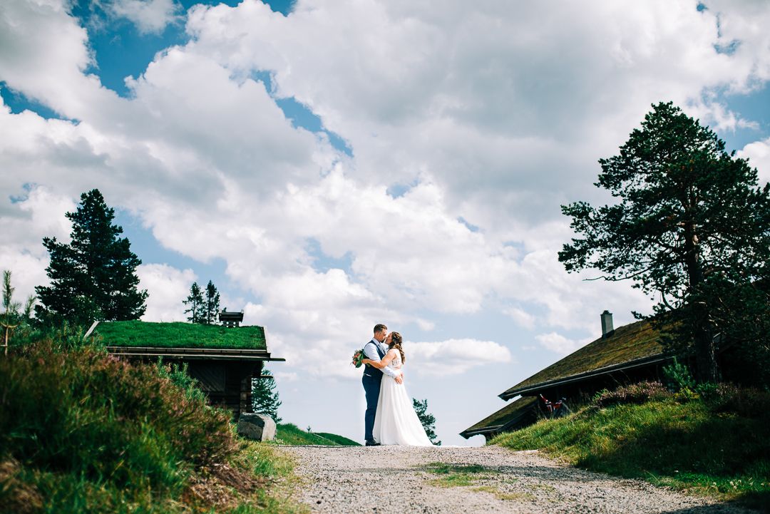 Wedding couple poses on gravel path between traditional Norwegian mountain cabins.