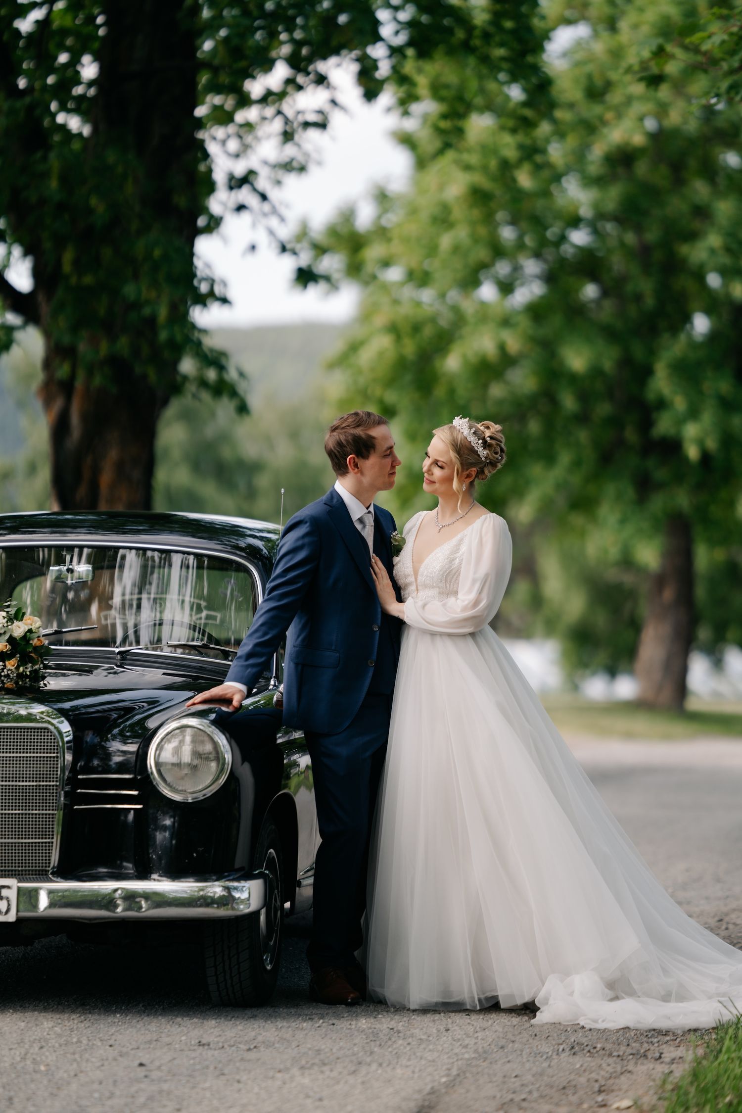 Bride and groom pose beside a classic black vintage car surrounded by lush green trees on their wedding day.