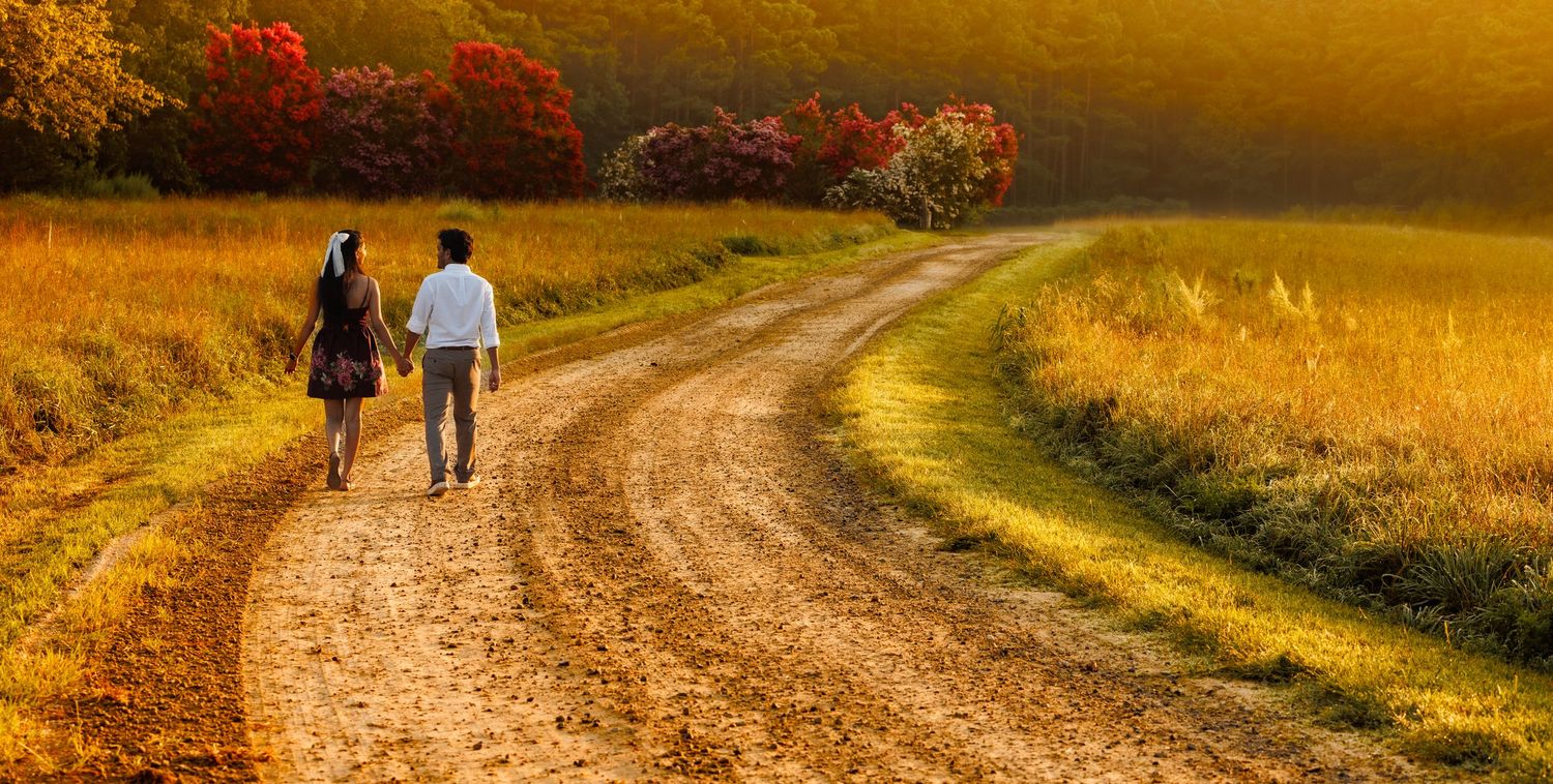 Couple walking down winding dirt path through golden field during sunset.