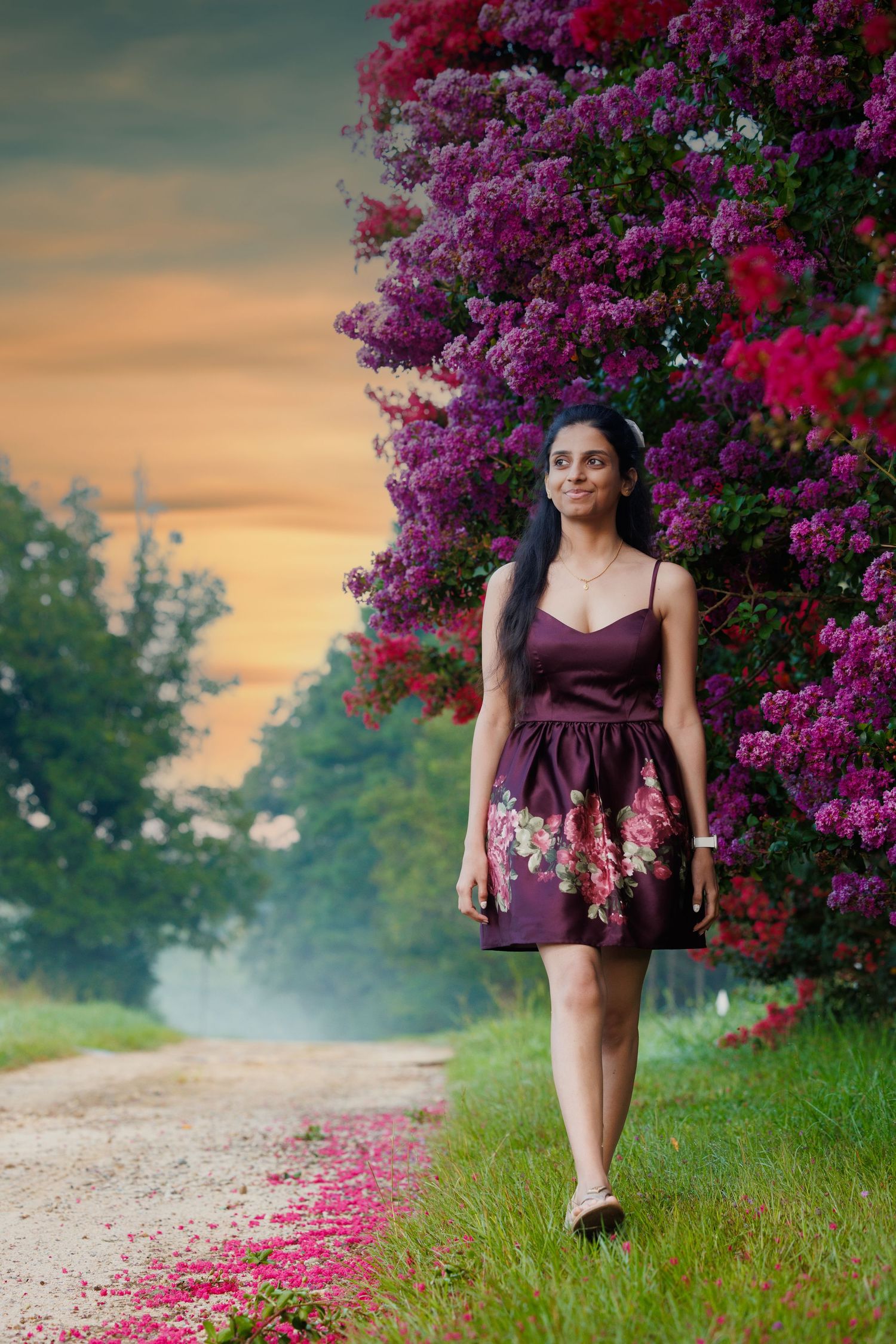 Woman in purple dress walking along path with blooming bougainvillea and sunset sky.