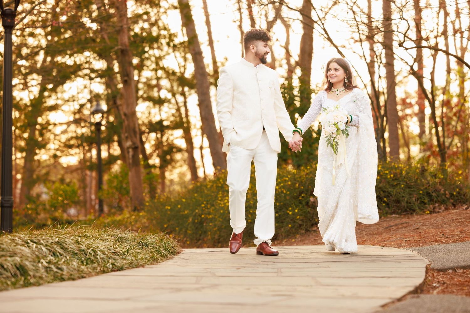 A couple in matching white outfits walking hand in hand on a wooden path during sunset.