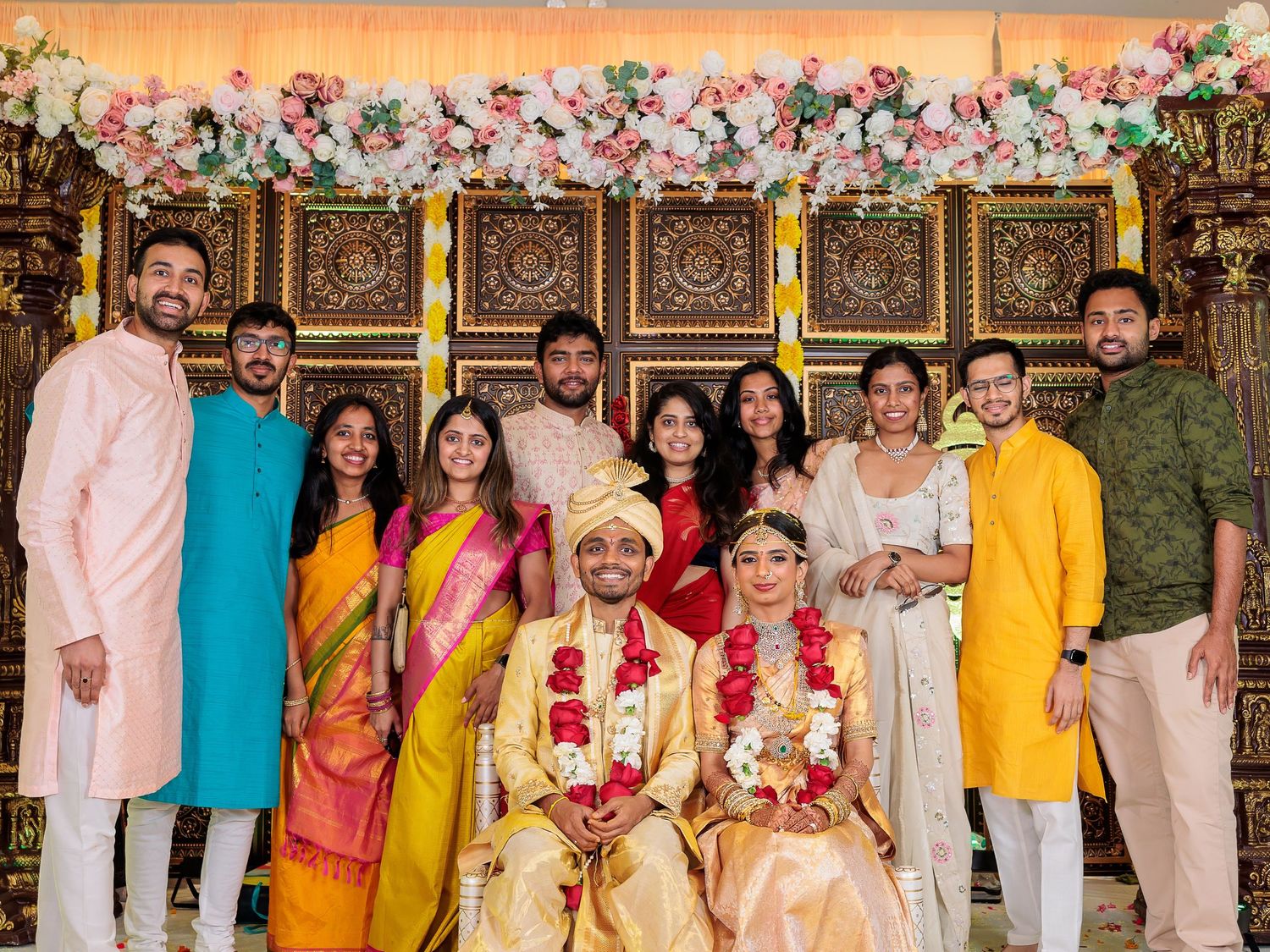 A family wedding photo showing traditional Indian attire and decorative floral arrangements in an ornate setting.