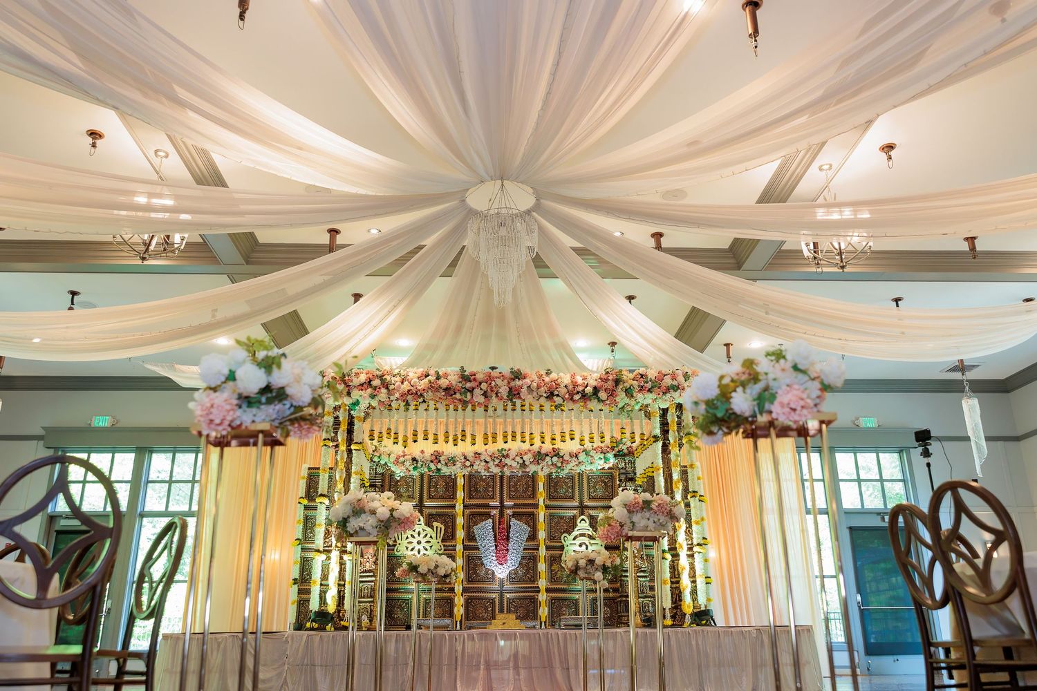 Wedding mandap decorated with white drapes and floral arrangements under elegant chandelier lighting.
