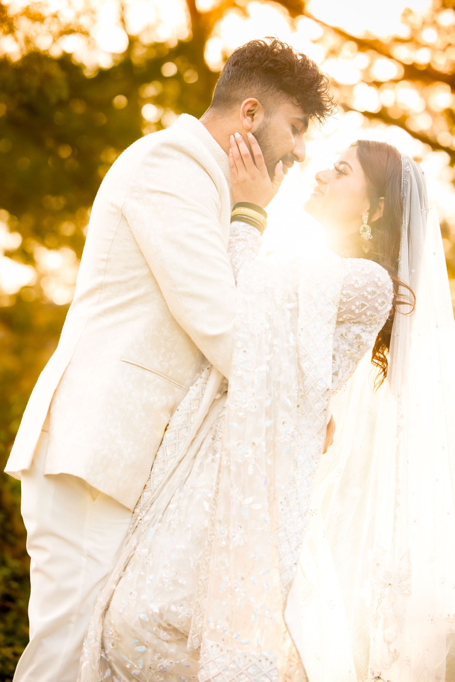A tender moment between newlyweds in white attire backlit by golden sunlight.