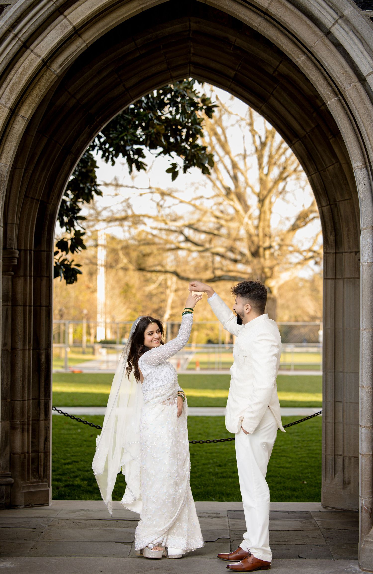 A couple in white wedding attire dance under a stone archway during a romantic outdoor photoshoot.