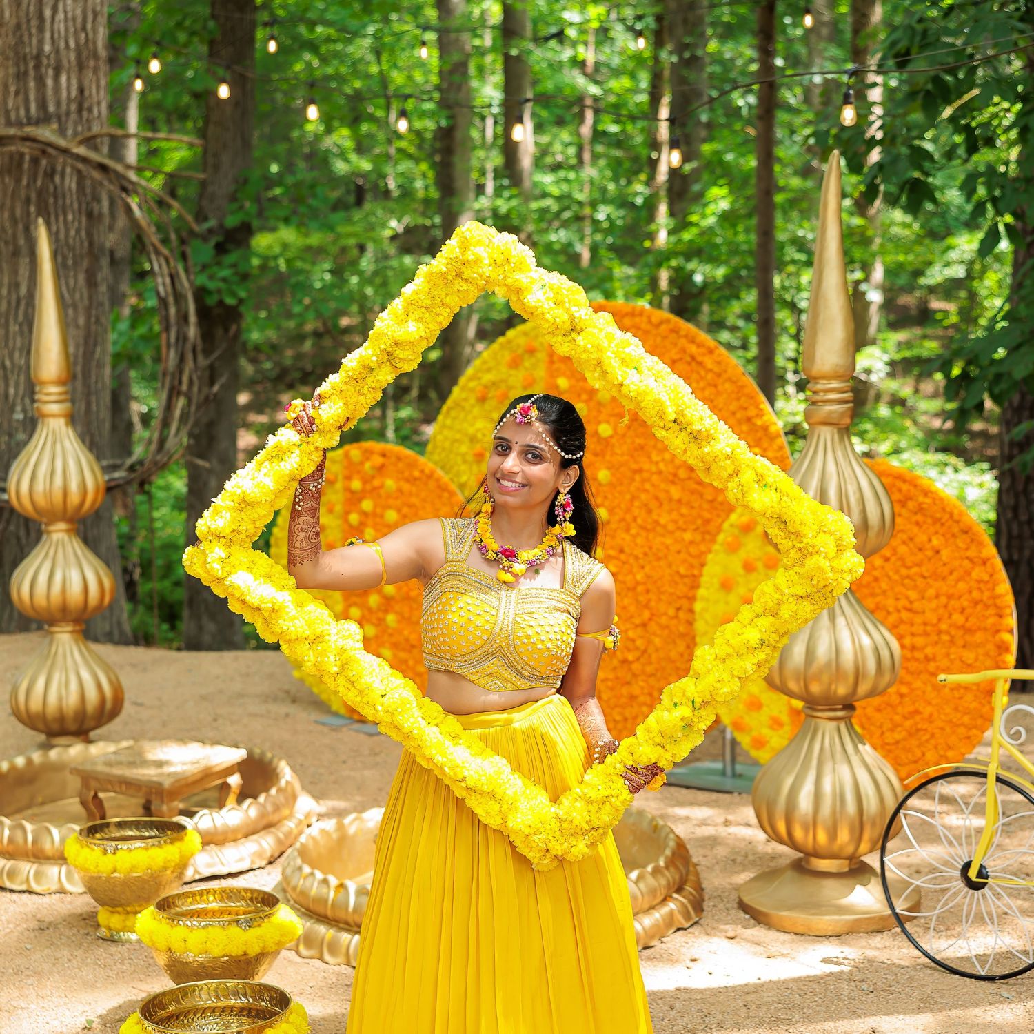 Traditional Indian wedding decor features a vibrant yellow floral frame with gold accents in an outdoor garden setting.