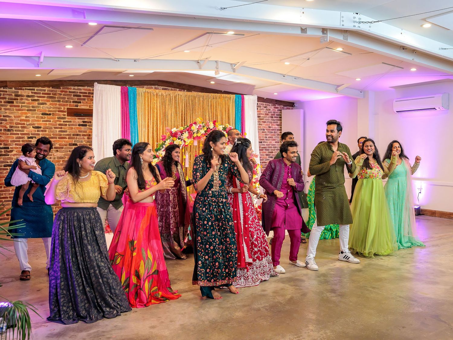 Wedding guests dancing in colorful traditional Indian attire at indoor celebration.