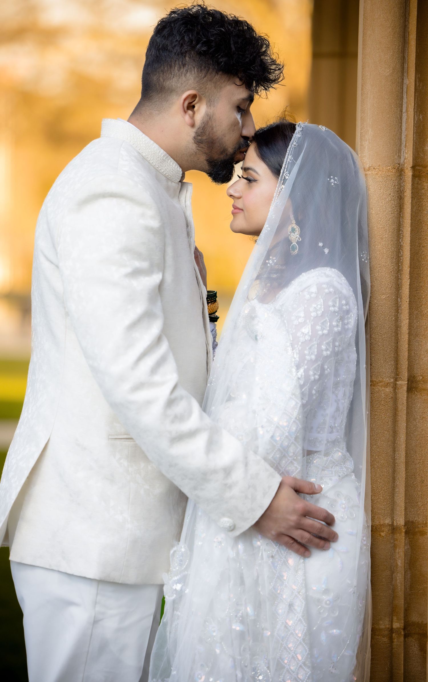 An intimate wedding portrait of a couple in white attire sharing a tender moment against a stone wall.