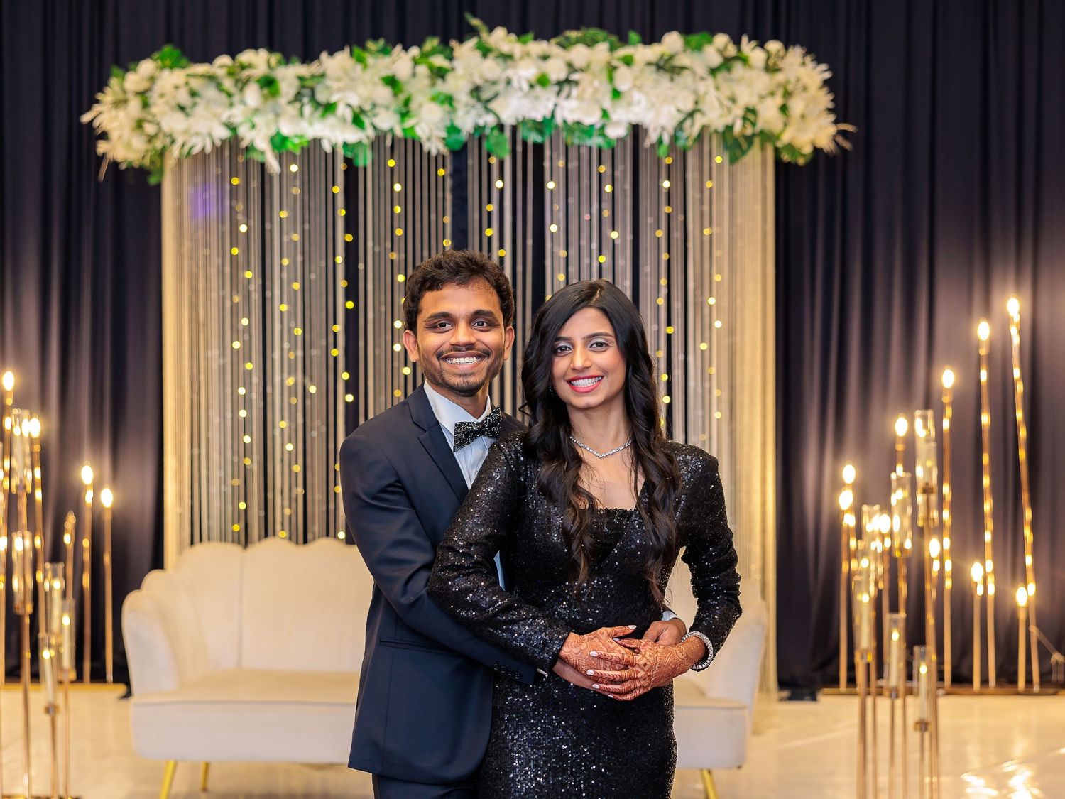 Couple smiling in formal attire against illuminated floral backdrop at evening event.