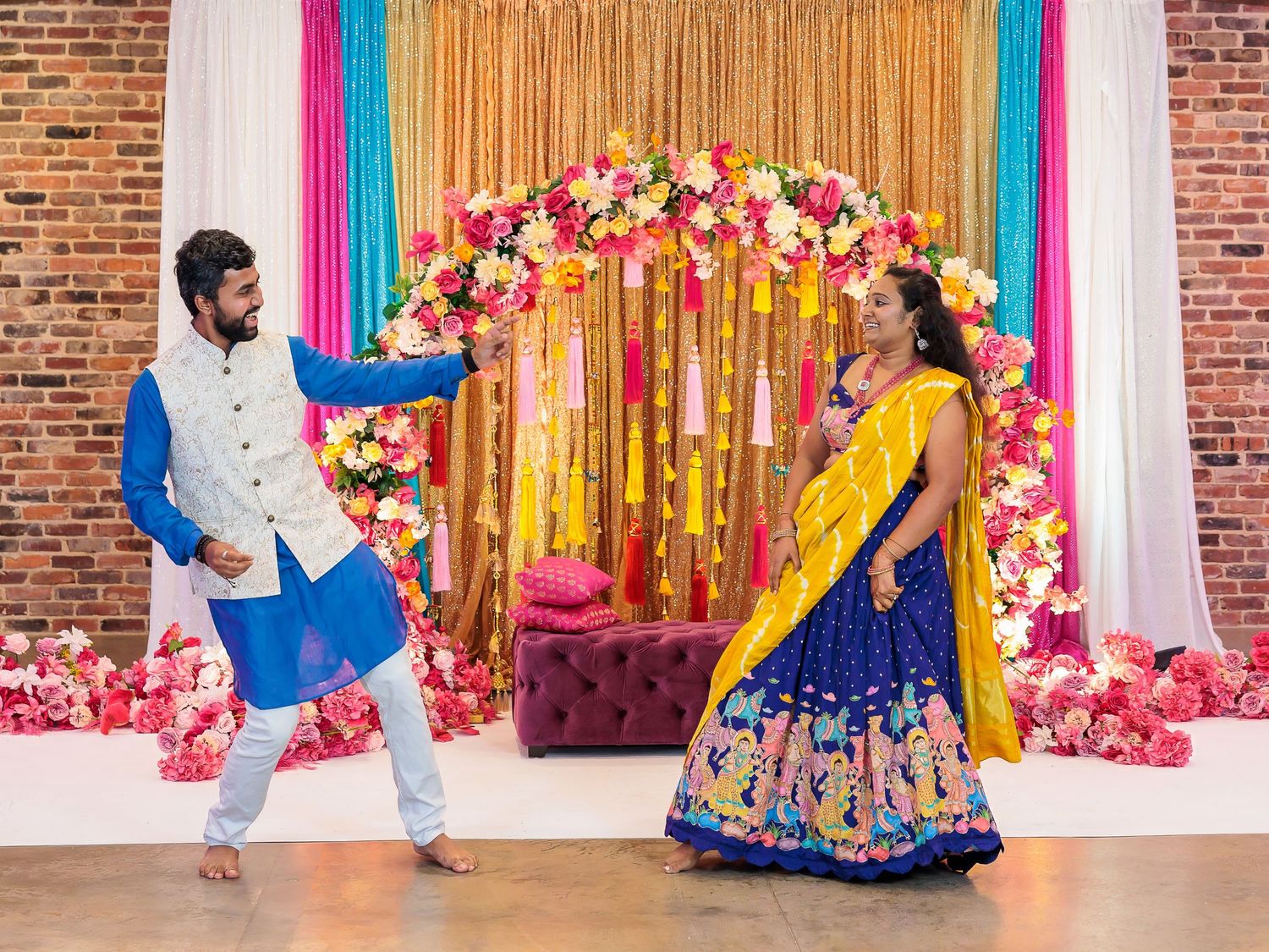 Colorful wedding celebration with floral arch and multicolored drapes against brick wall backdrop.
