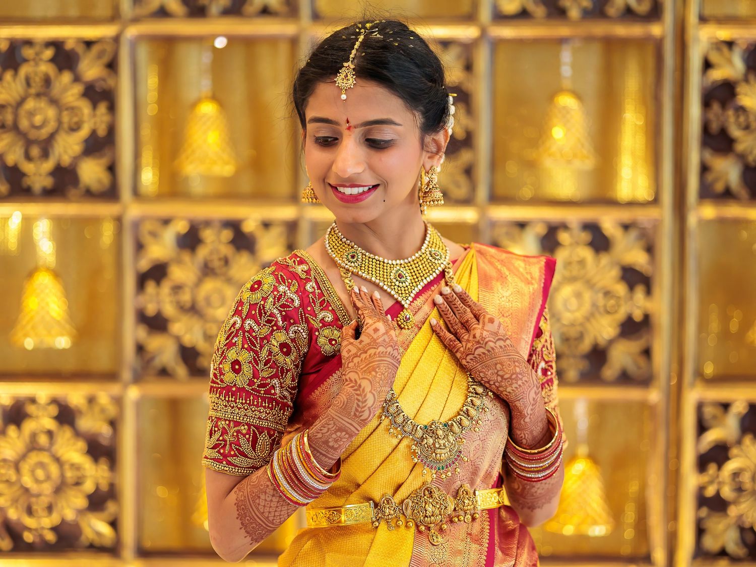 Traditional South Indian bridal portrait with gold jewelry against ornate golden bell backdrop.