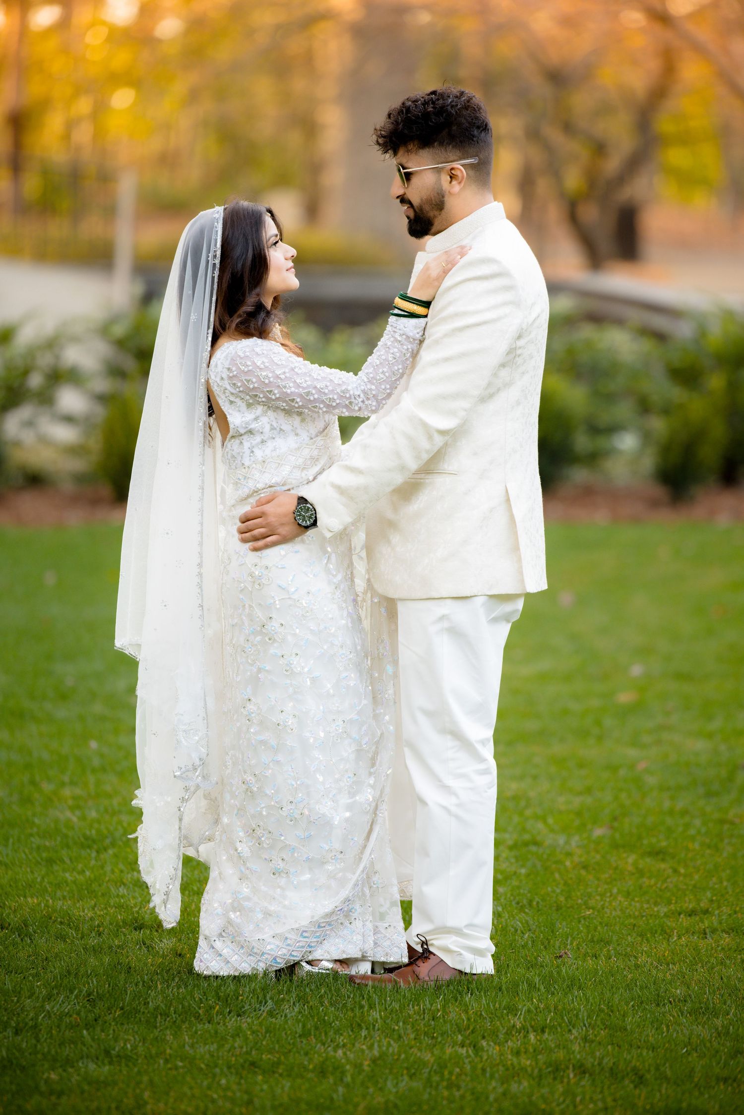 A romantic wedding portrait of a couple in white attire embracing on a grassy lawn during autumn.