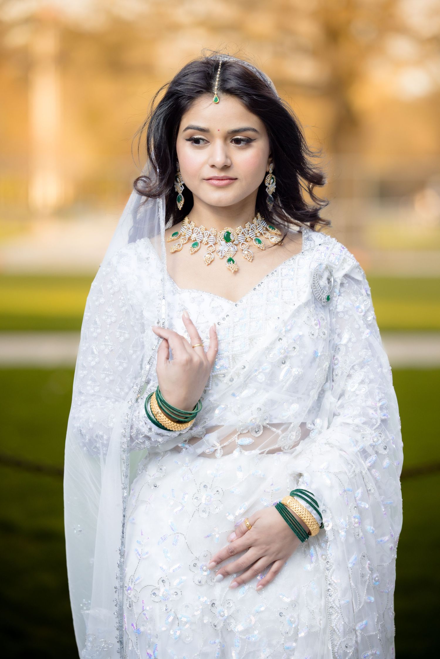 A bride in white traditional attire wearing emerald and crystal jewelry with flowing veil.
