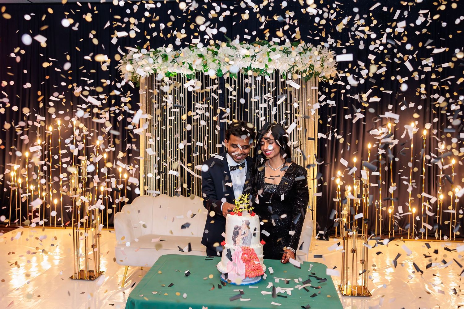Couple cutting cake under floral arch while confetti falls at celebration event.