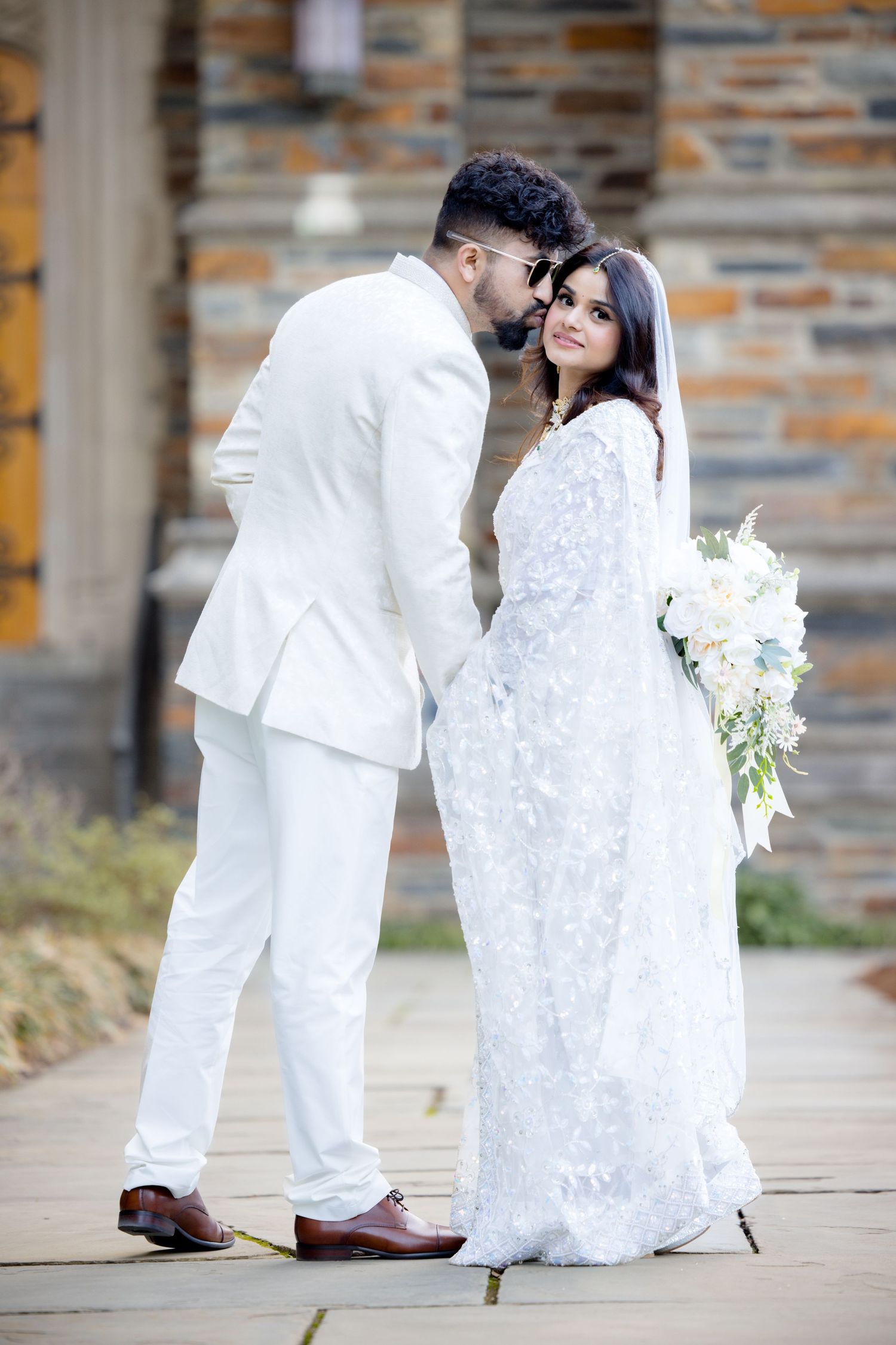 A romantic wedding portrait of a couple in all-white outfits standing by a stone wall.
