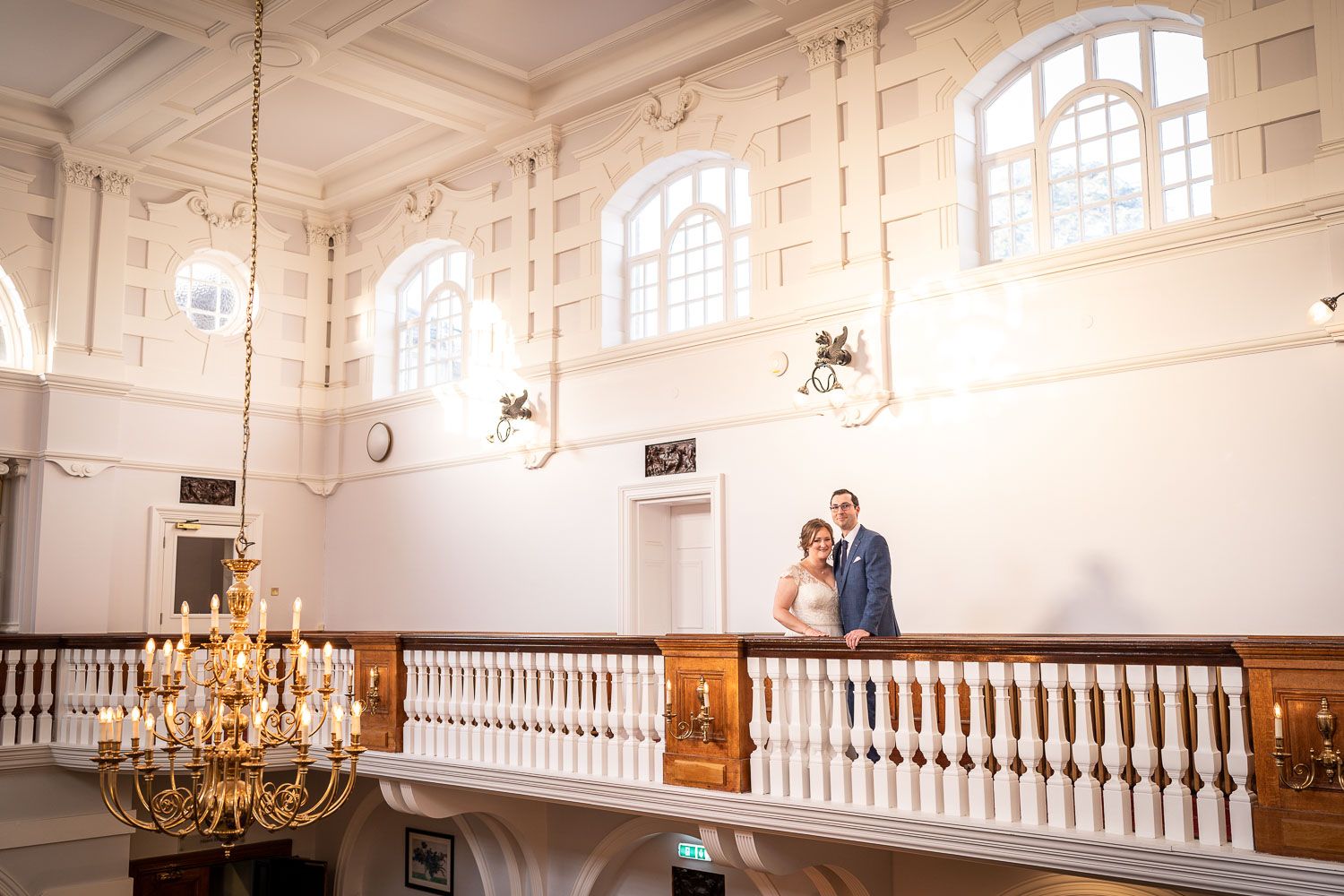 A couple stands together in an elegant white interior space with arched windows and a grand crystal chandelier.