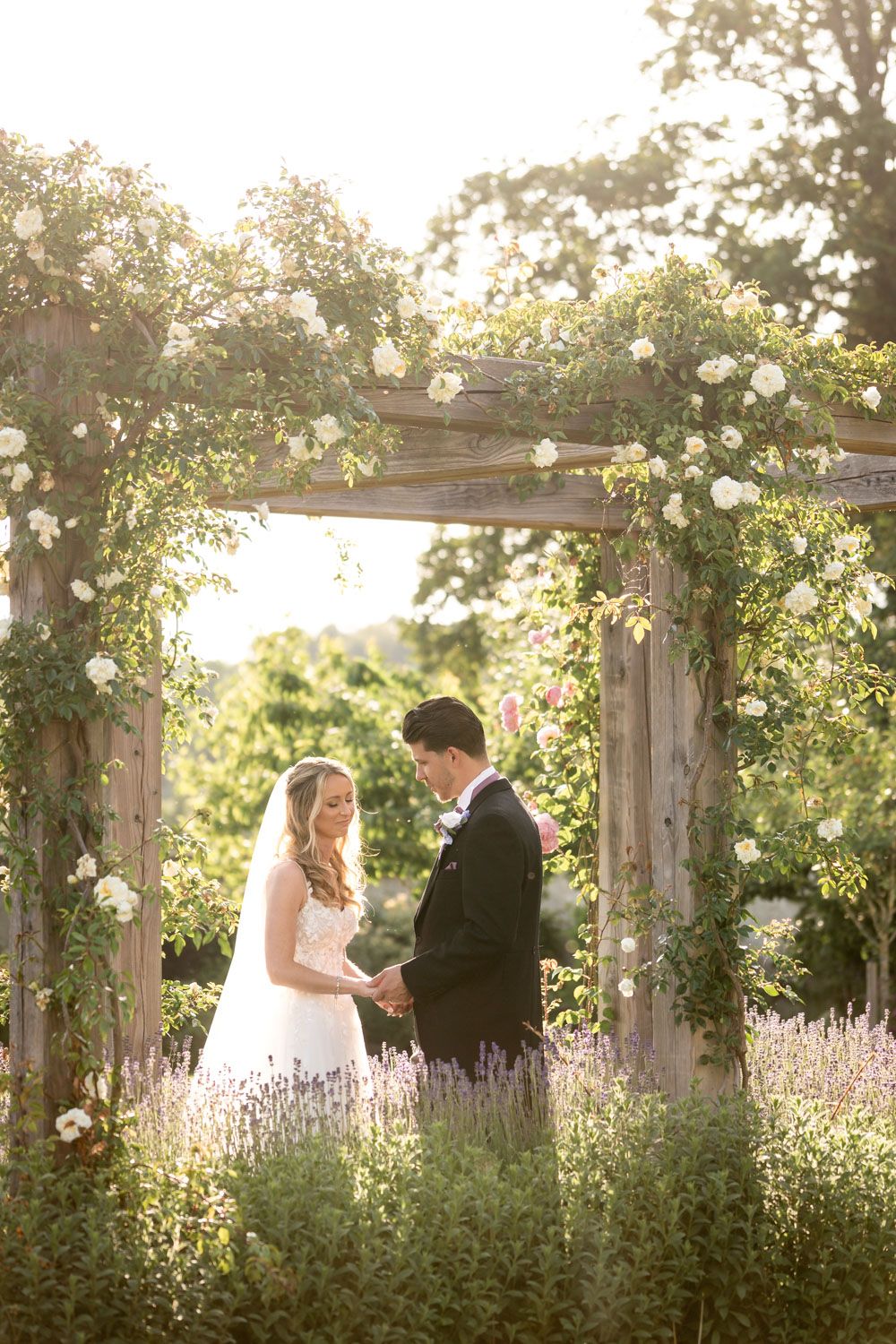A romantic garden wedding scene with a couple holding hands under a flower-covered pergola at golden hour.