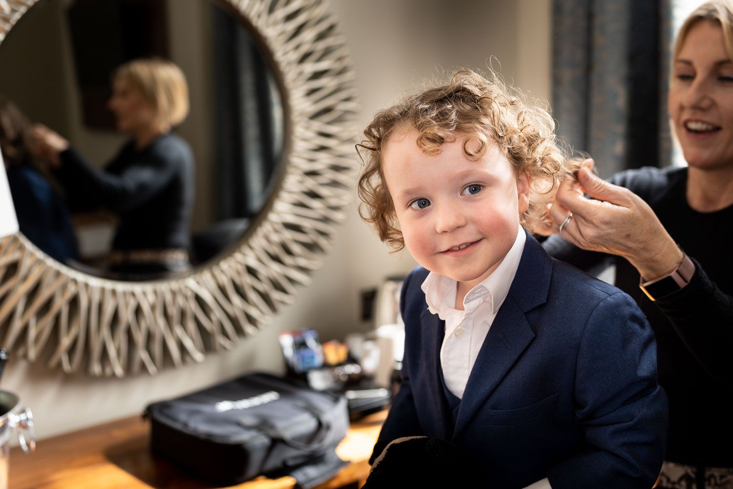 A child in a navy suit jacket and white shirt smiles while getting styled at a hair salon.