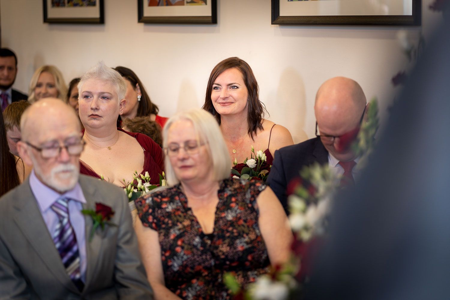 Wedding guests smile during an intimate indoor ceremony with decorative flowers and framed artwork on the walls.