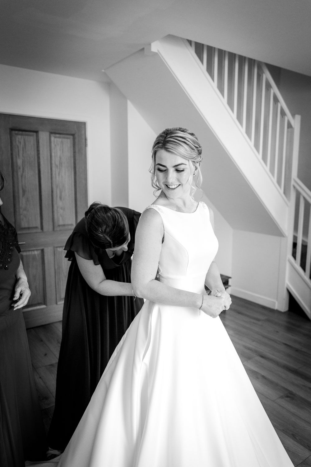 Black and white photo of a bride smiling as her bridesmaid fastens her elegant wedding gown before the ceremony.