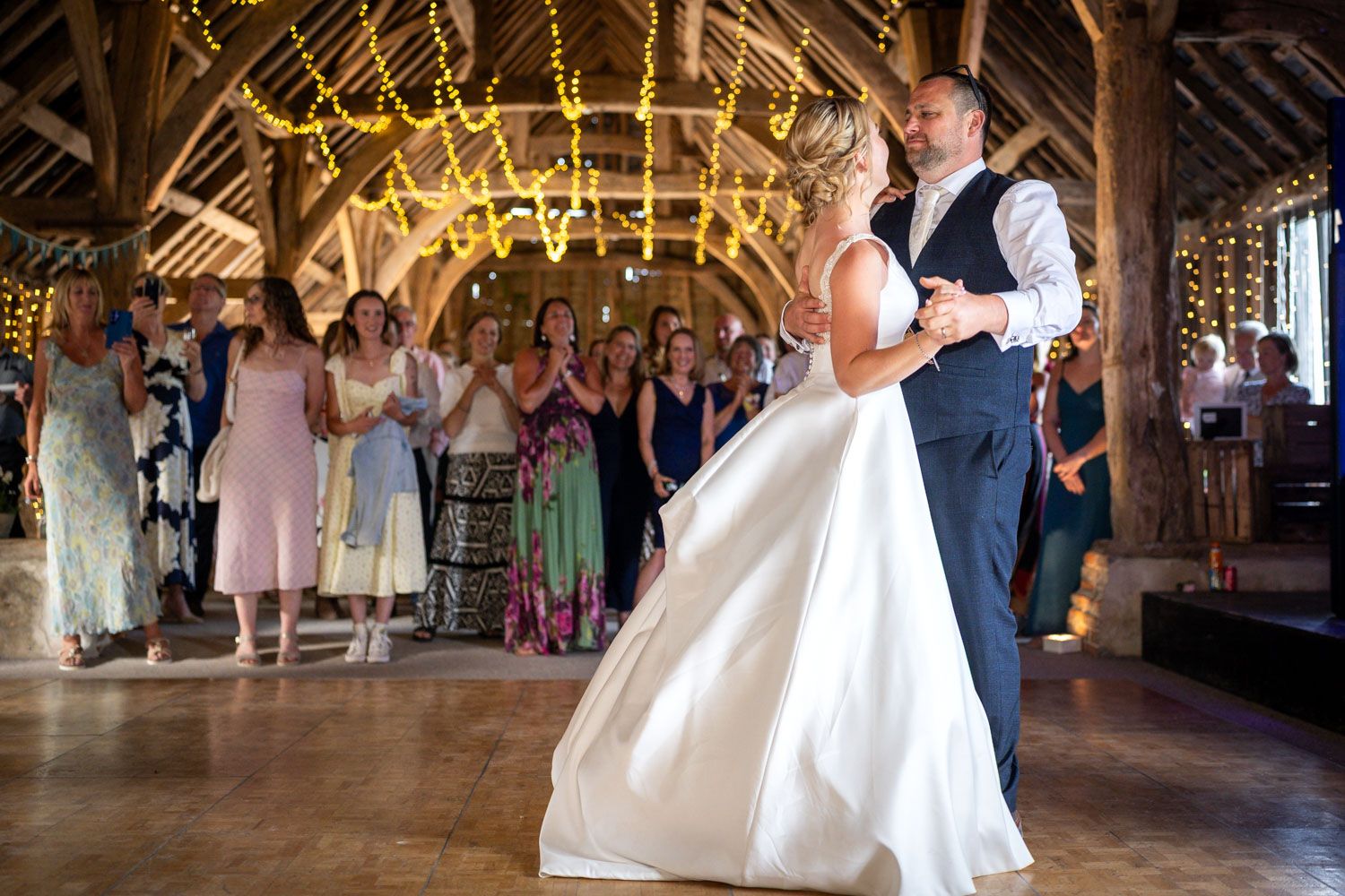 Bride and groom share first dance in rustic barn wedding venue with fairy lights and guests applauding in background.