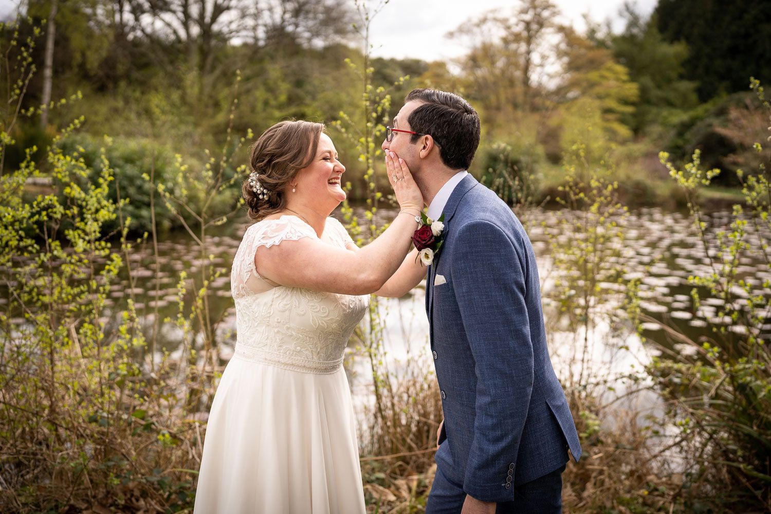 A romantic moment between a couple in formal wedding attire by a scenic pond with lily pads and greenery.
