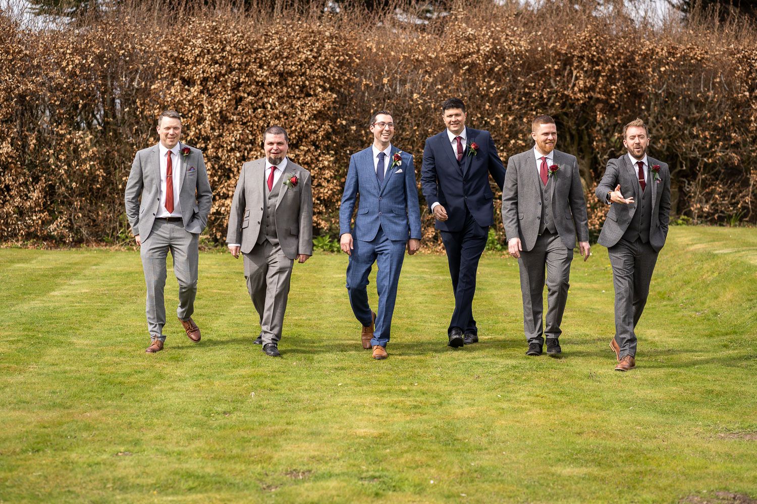Six groomsmen in tailored suits walking together across a green lawn.