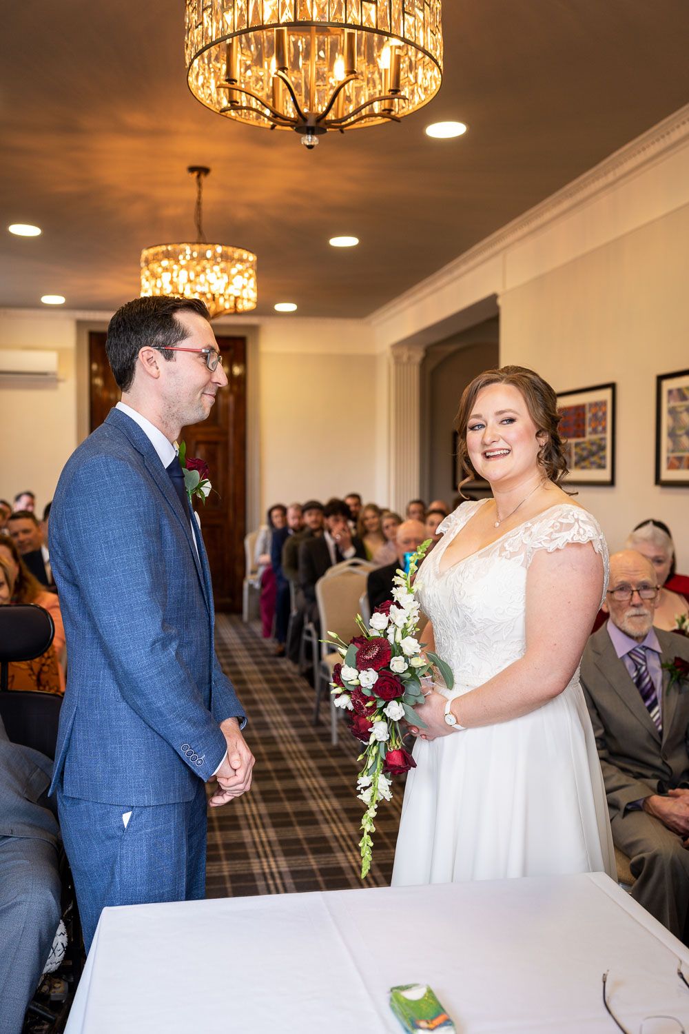 A wedding ceremony in an elegant room with crystal chandeliers as a couple stands at the front in formal attire.