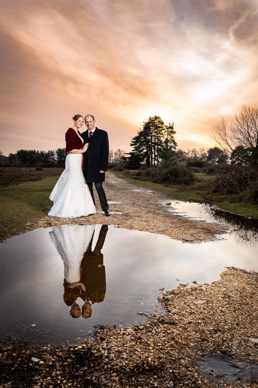 A couple in wedding attire stands by a puddle reflecting their silhouettes against a dramatic sunset sky over a rural path.