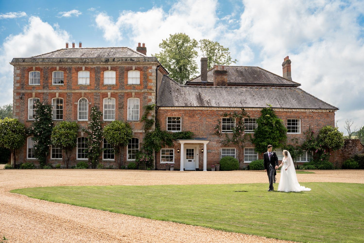 Grand brick manor house wedding venue with manicured lawn under blue summer sky.