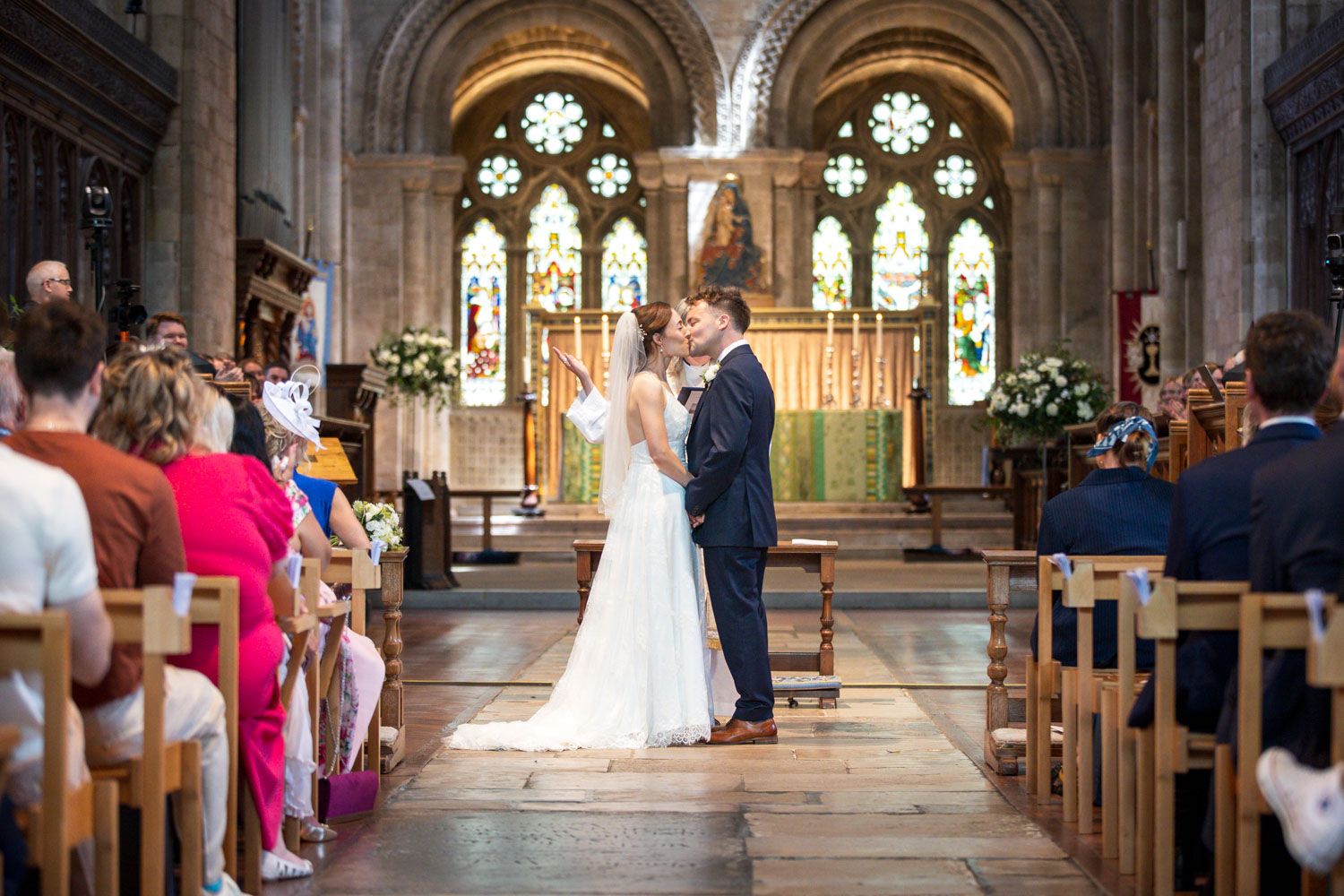 A traditional church wedding ceremony takes place beneath gothic arched windows and stained glass in a historic stone chapel.
