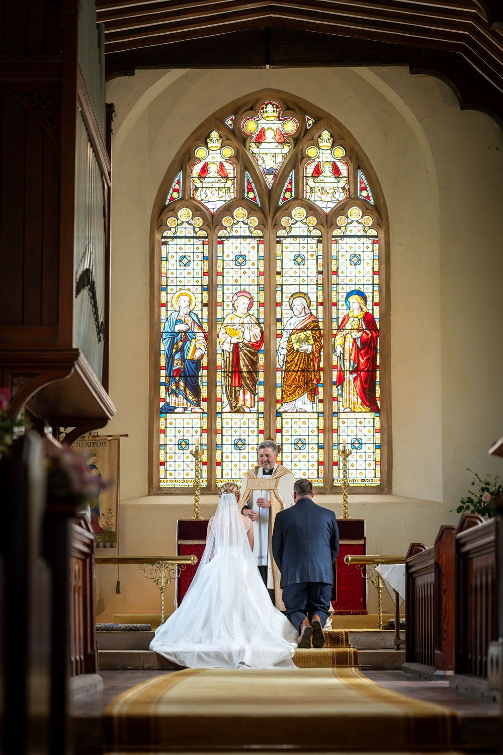 Bride and groom kneeling at church altar before officiant, with colorful stained glass window backdrop.