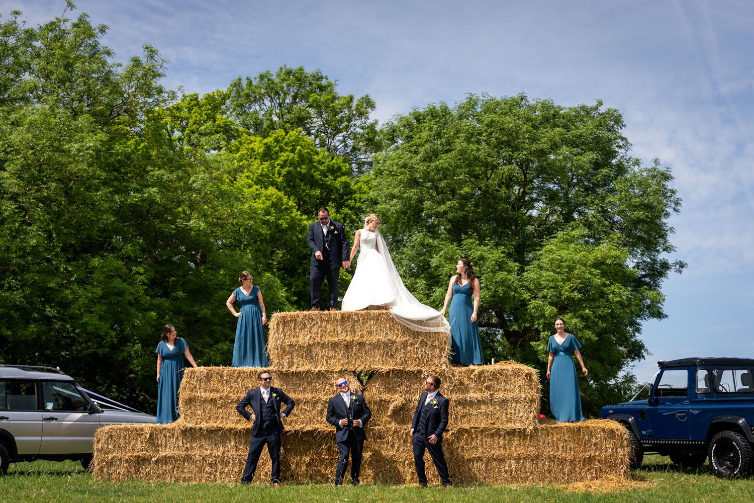Wedding party posed on hay bales outdoors, bride and groom at top, bridesmaids in teal gowns and groomsmen below.