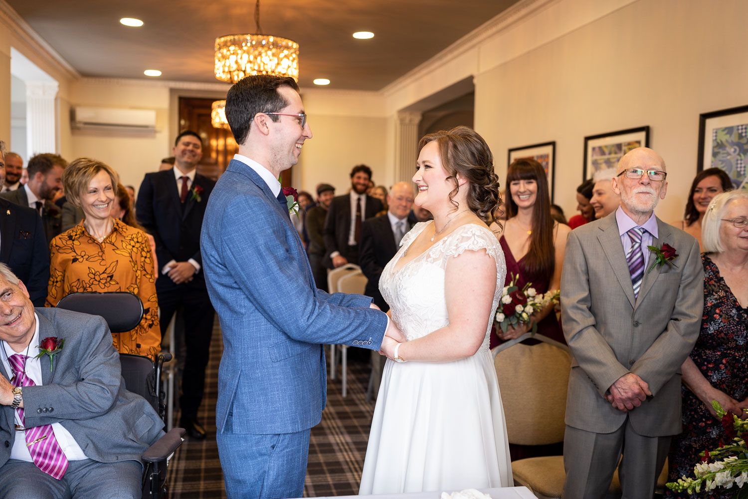 A couple exchanges vows during their indoor wedding ceremony surrounded by smiling guests and decorative lighting.