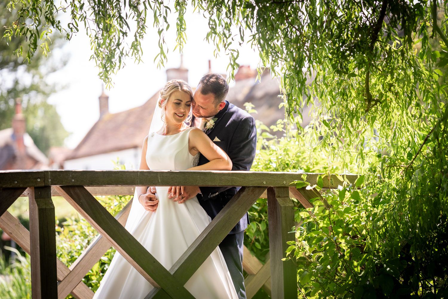 Bride and groom embracing on a wooden bridge surrounded by lush weeping willow trees at an English countryside wedding venue.