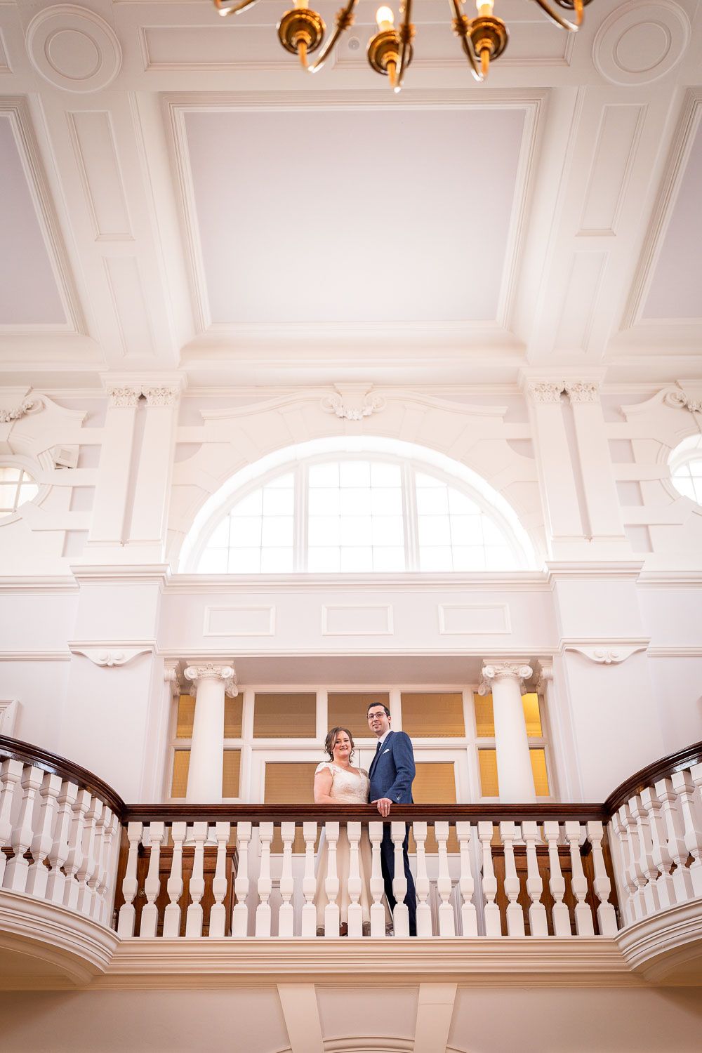 Elegant couple stands on ornate white interior balcony with grand arched window and decorative moldings.
