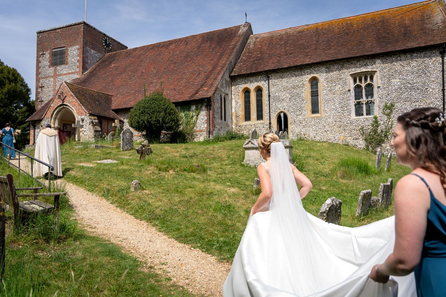 Bride in white gown and veil walks toward a historic stone church, bridesmaid carrying her train on a sunny day.