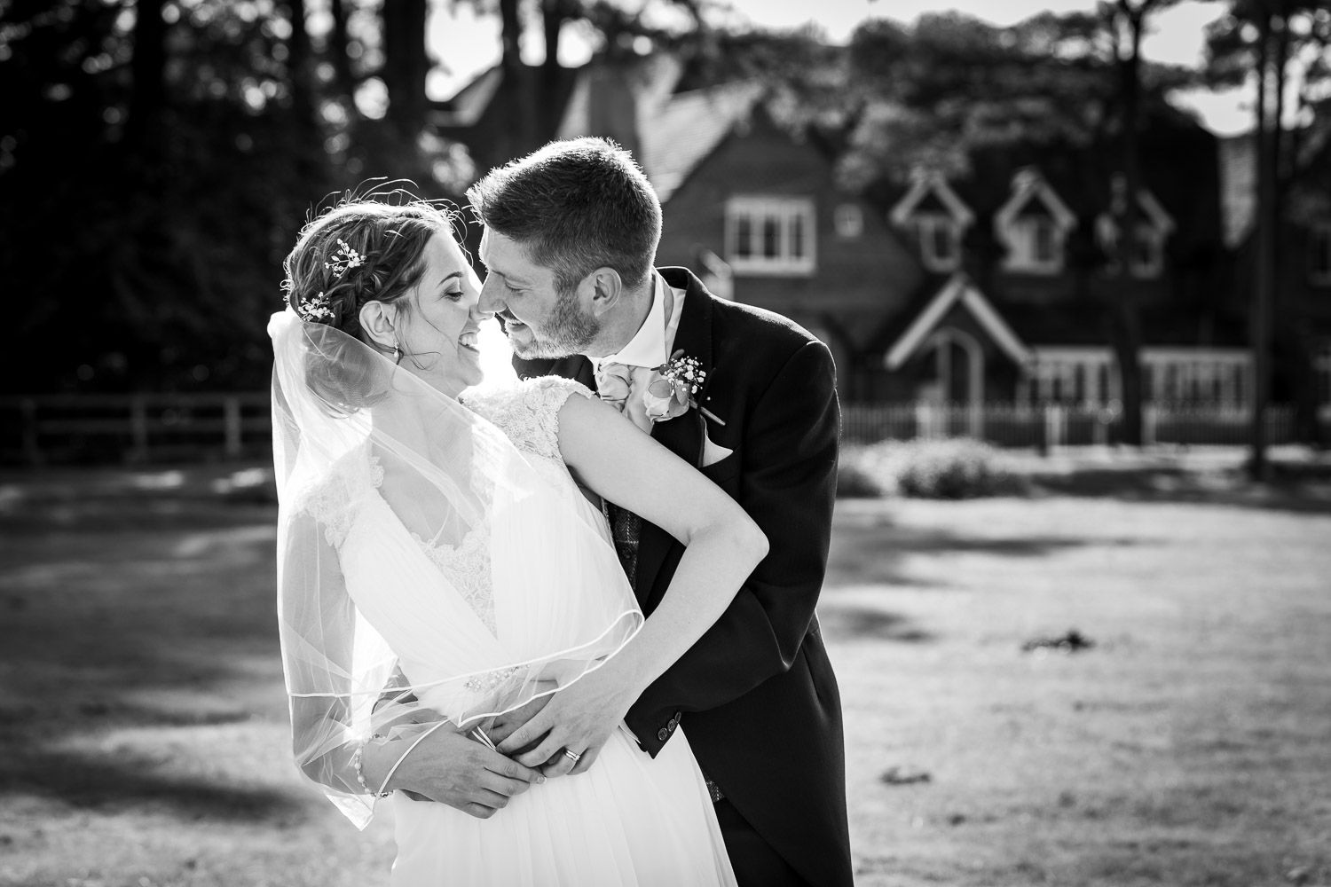 A romantic black and white wedding moment captured outdoors with a couple embracing at a country venue with houses in background.