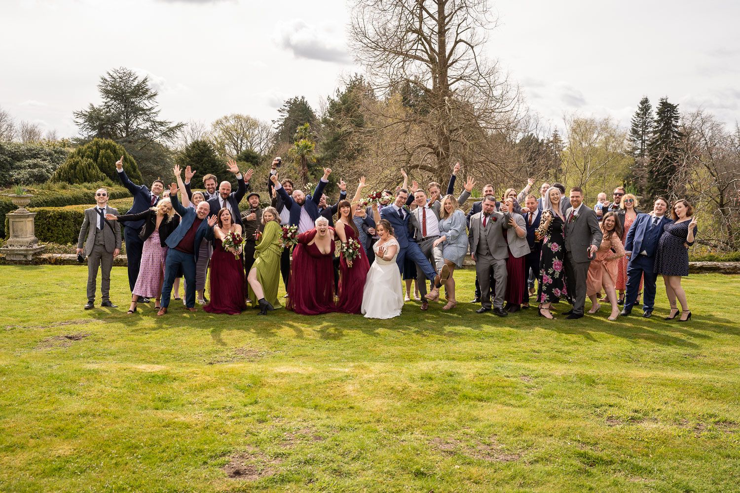 Large wedding group photo with guests in burgundy and formal attire gathered on green lawn.