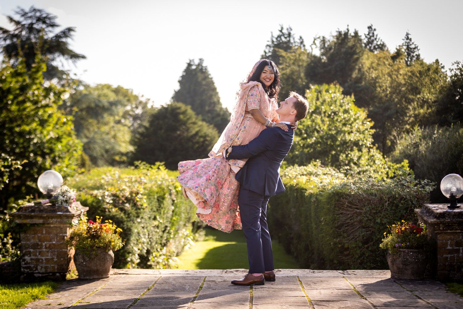 A couple in formal attire dance in a sunlit garden with ornate potted flowers and greenery.