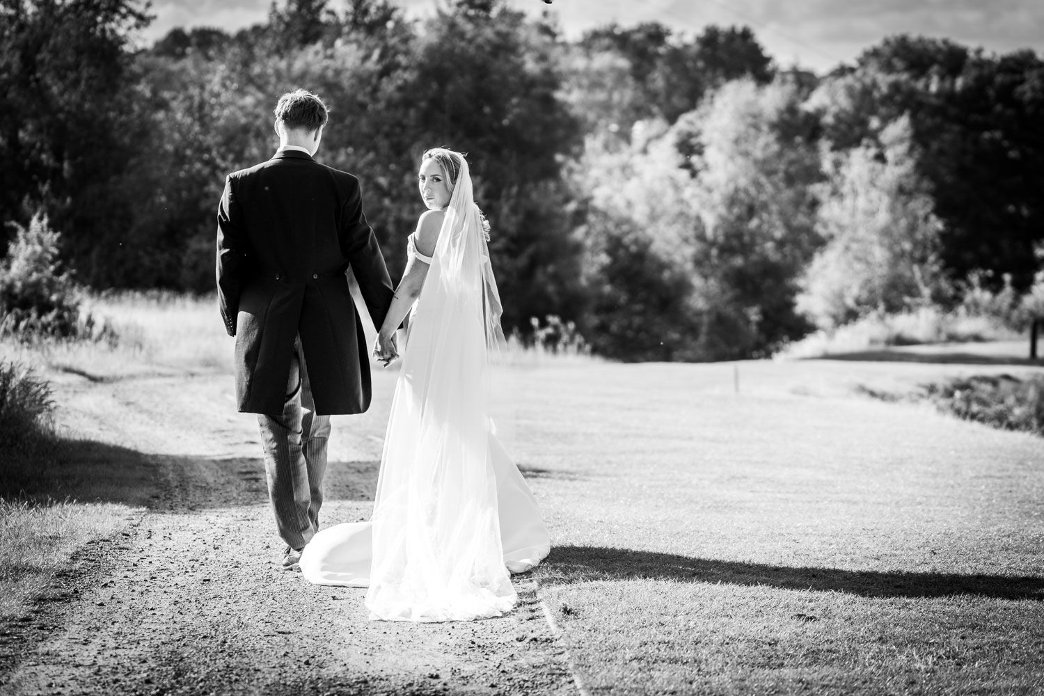 Elegant black and white photograph of a bride and groom walking together on a winding path surrounded by trees.