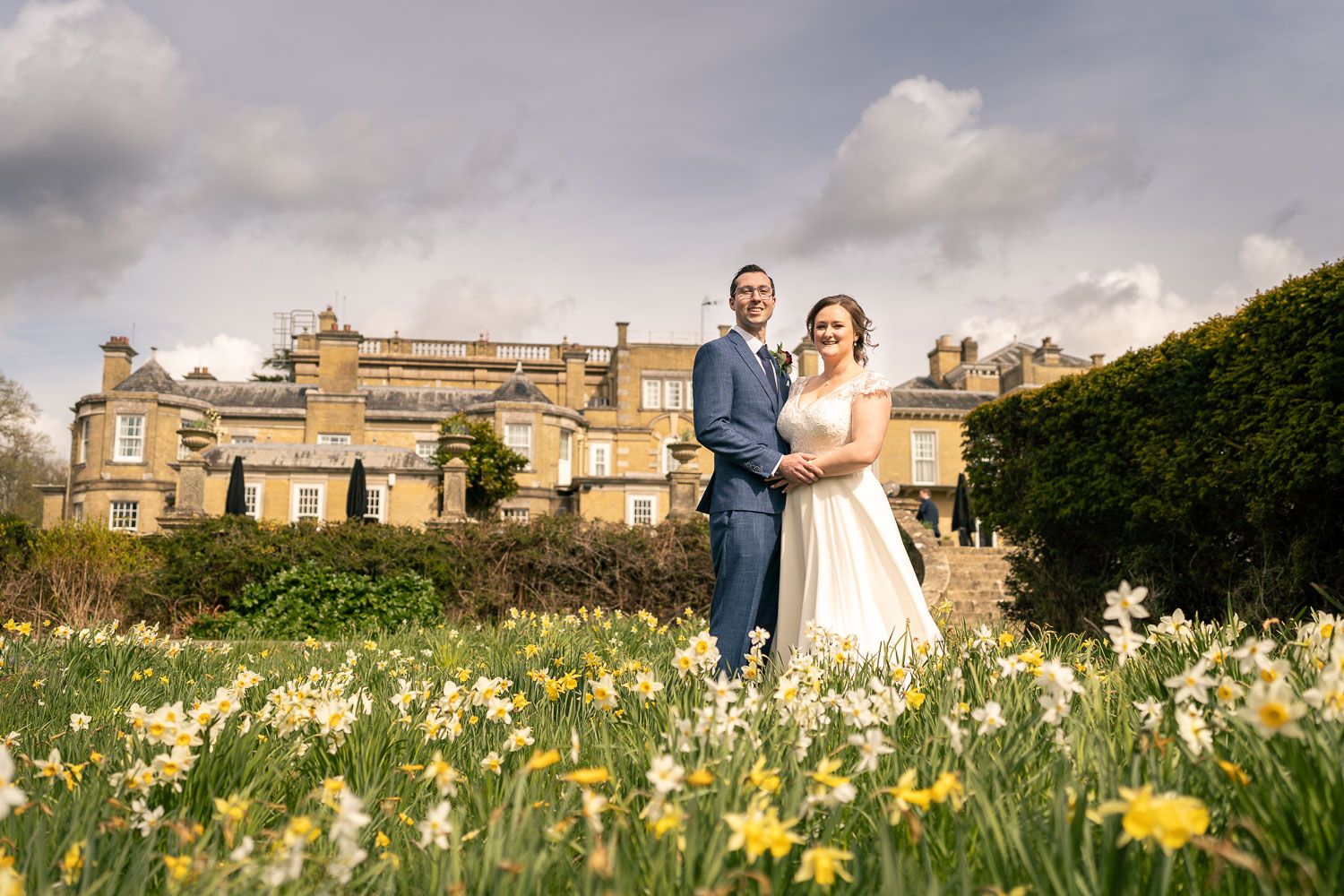 A couple stands in a beautiful garden filled with blooming daffodils in front of a grand historic mansion.