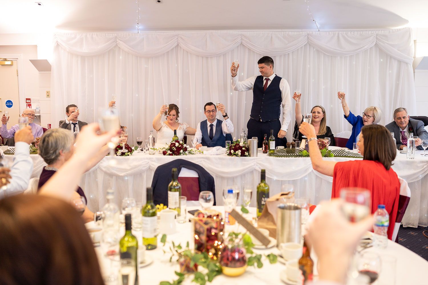 A waiter stands on a chair making an announcement to guests at an elegant wedding reception with white draped decor.