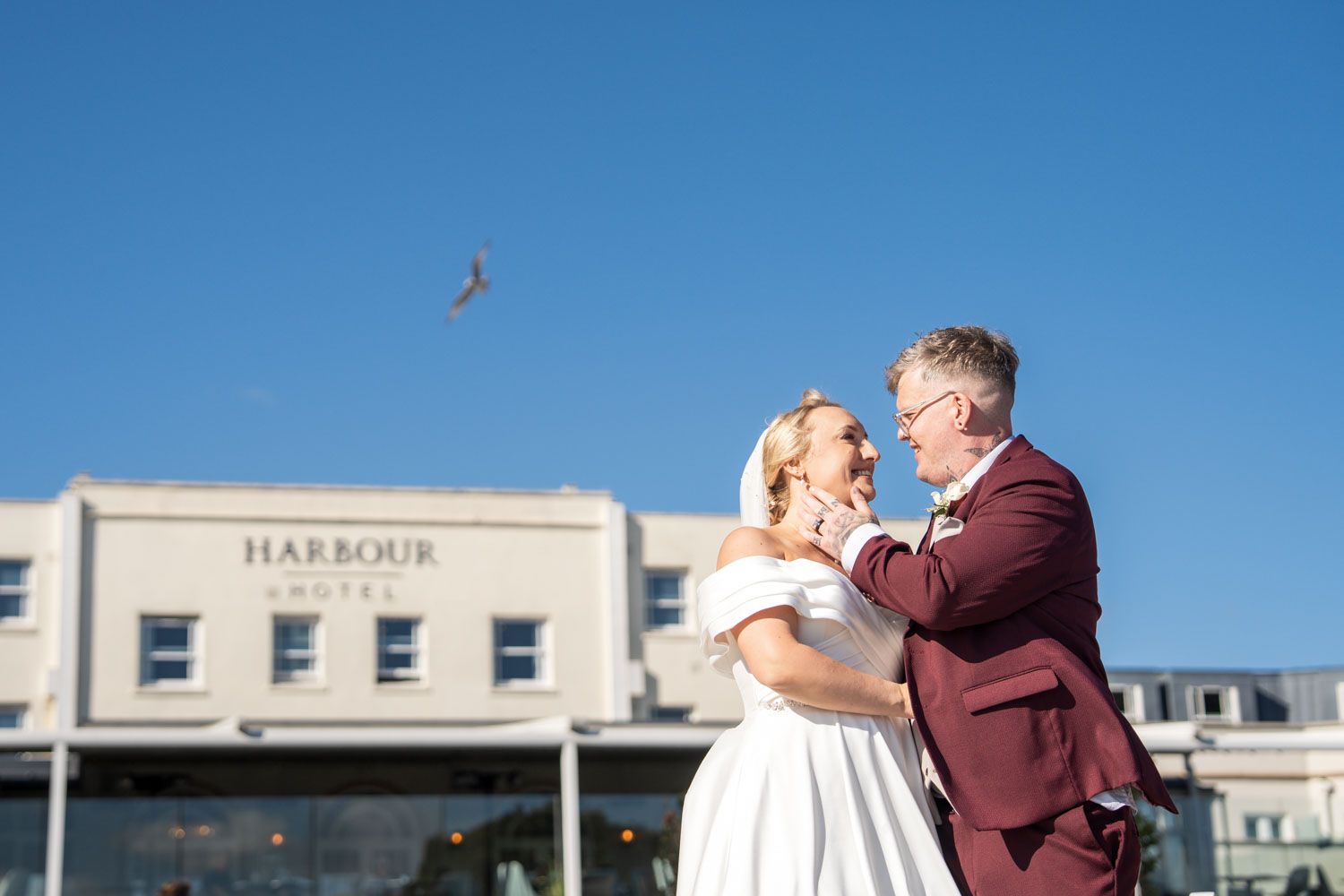 A couple in wedding attire embrace in front of a building labeled 'Harbour' on a sunny day with blue skies.