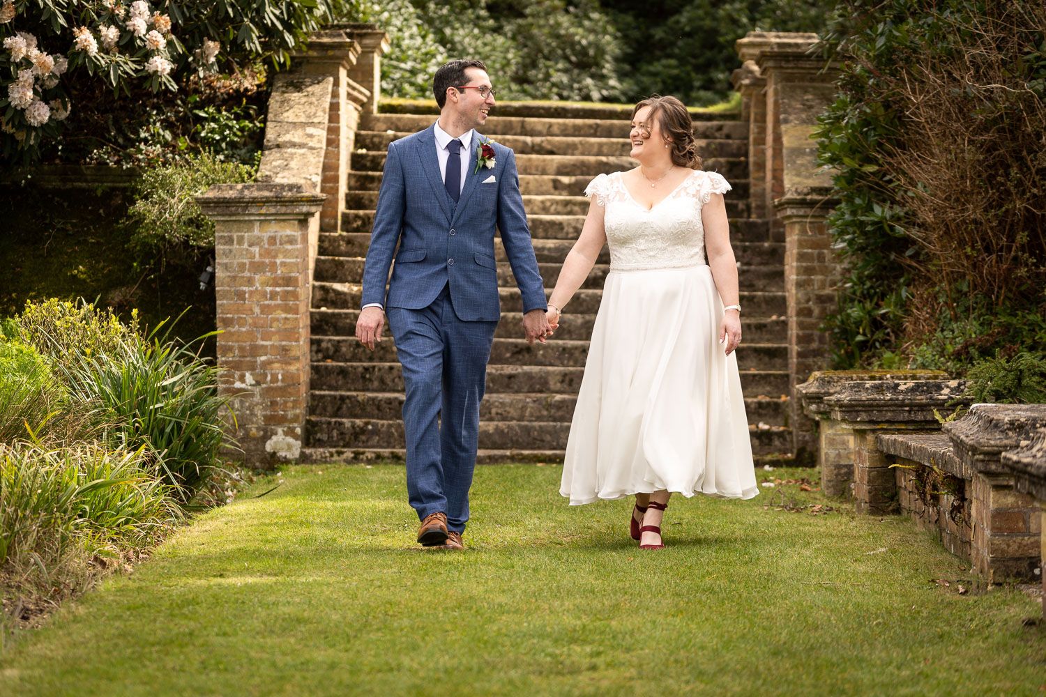 A couple walks down stone garden steps holding hands, with the individual in a navy suit leading the way.