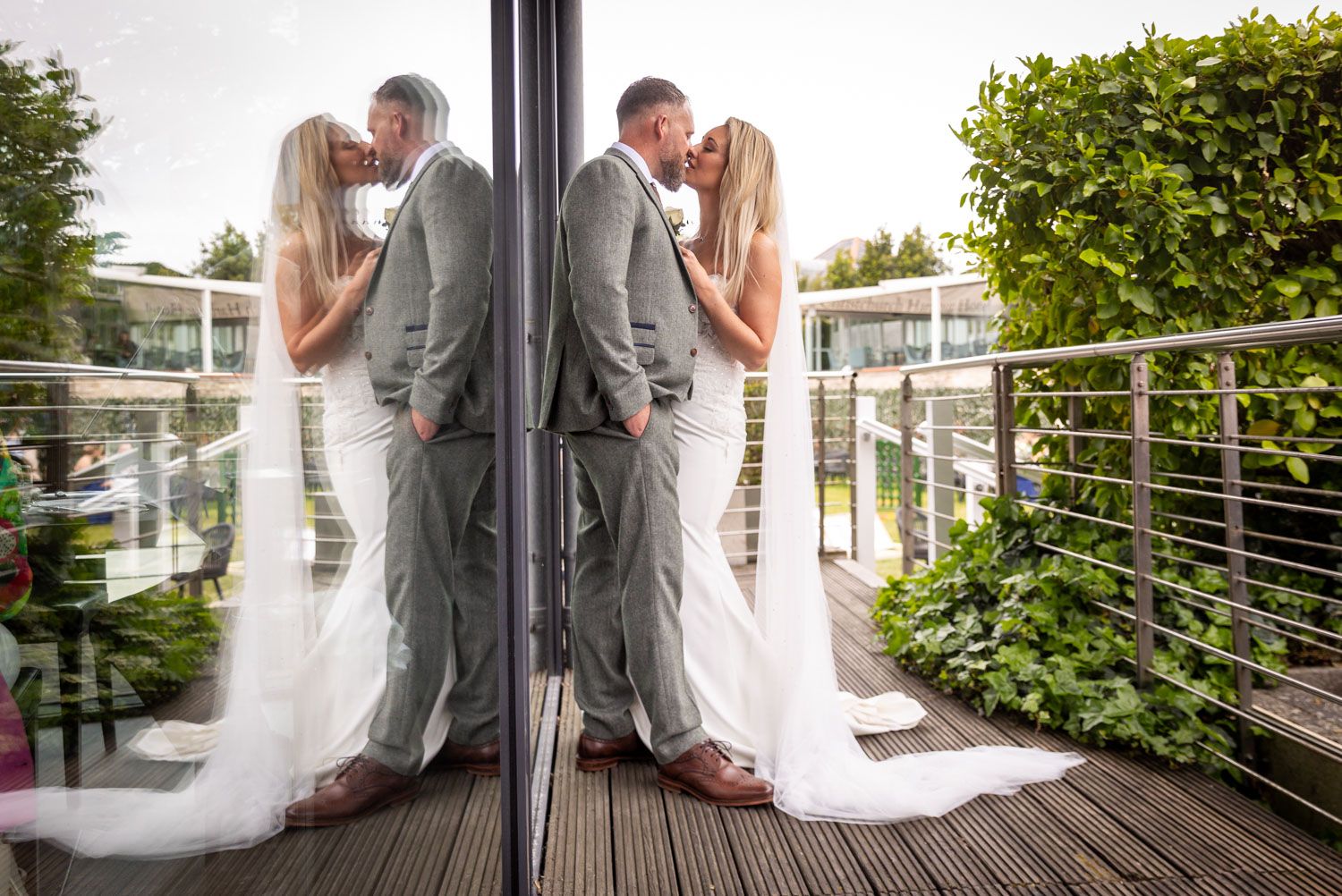 A bride and groom share a kiss on a wooden deck, beautifully reflected in a glass panel at the Harbour Hotel Christchurch