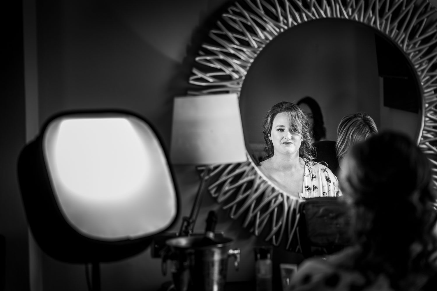 Black and white reflection in vanity mirror showing figure illuminated by lamp light in moody setting.